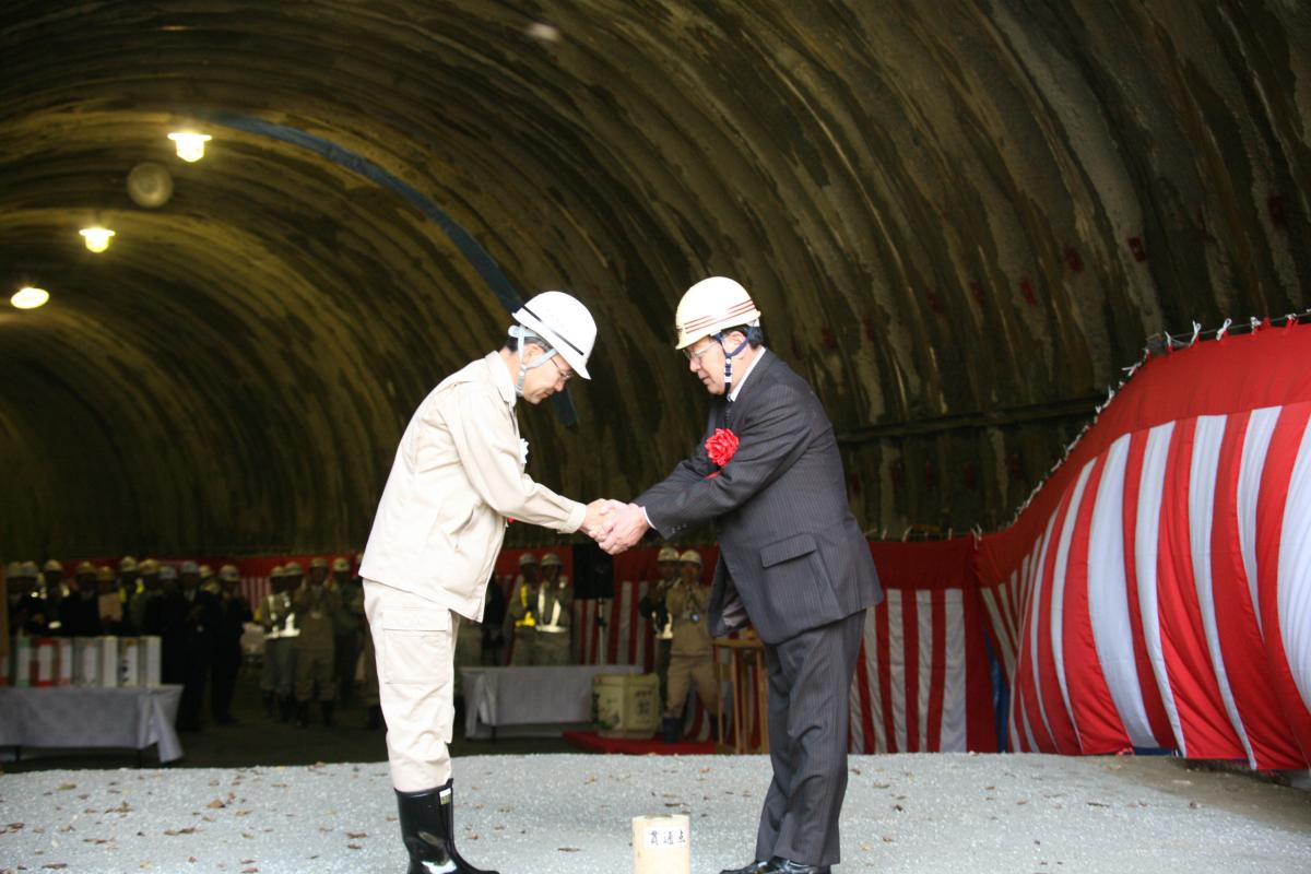 見事貫通したトンネル内で握手を交わす渡辺町長と有我相双建設事務所長