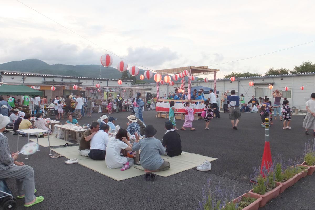 やぐらを囲み盆踊りを踊る子どもたちと、ござに座りおいしいものを食べながら夏祭りを楽しむ町民