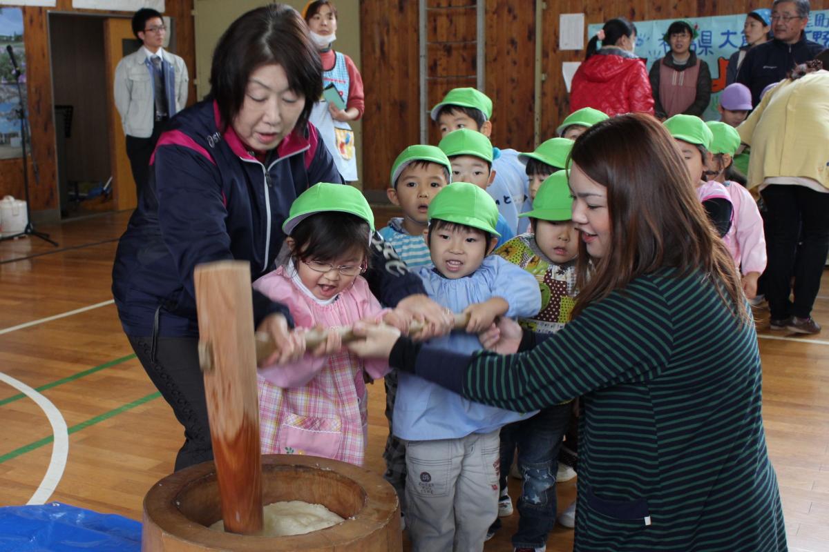 先生にも手伝ってもらいもちをつく園児と後ろから覗き込むようにもちつきの様子を見る園児