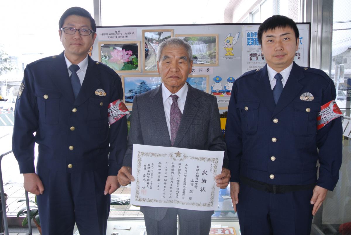 会津若松警察署の玄関に飾られた写真を前に感謝状を手にする山田さん