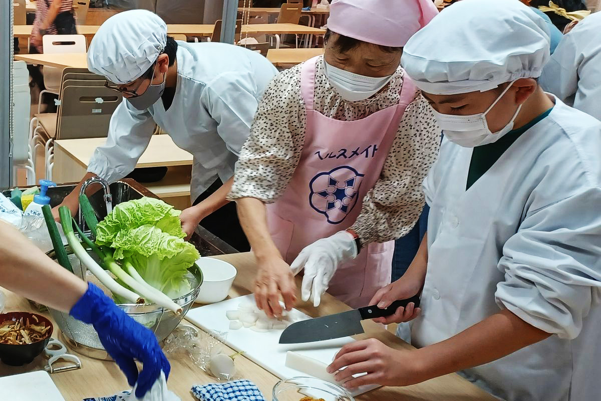 義務教育課程の子どもたちは2組に分かれ、おにぎりや豚汁、サツマイモのおふかしを作りました。家庭科室では豚汁作りが行われ、包丁を使う子どもたちは基本的な使い方や動かし方、材料の切り方などボランティアの方から付きっ切りで教えてもらいました。