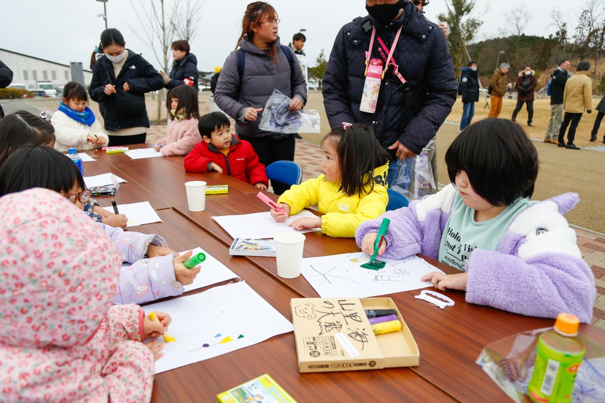 里がえりもちつき大会のイベントでは、餅つき体験の他に、お正月遊びとして「たこあげ」や巨大かるたを使った「人間カルタ」、三輪車で接戦を繰り広げる「おおくま競輪」が行われました。写真は、たこのデザインを描く子どもたちの様子です。