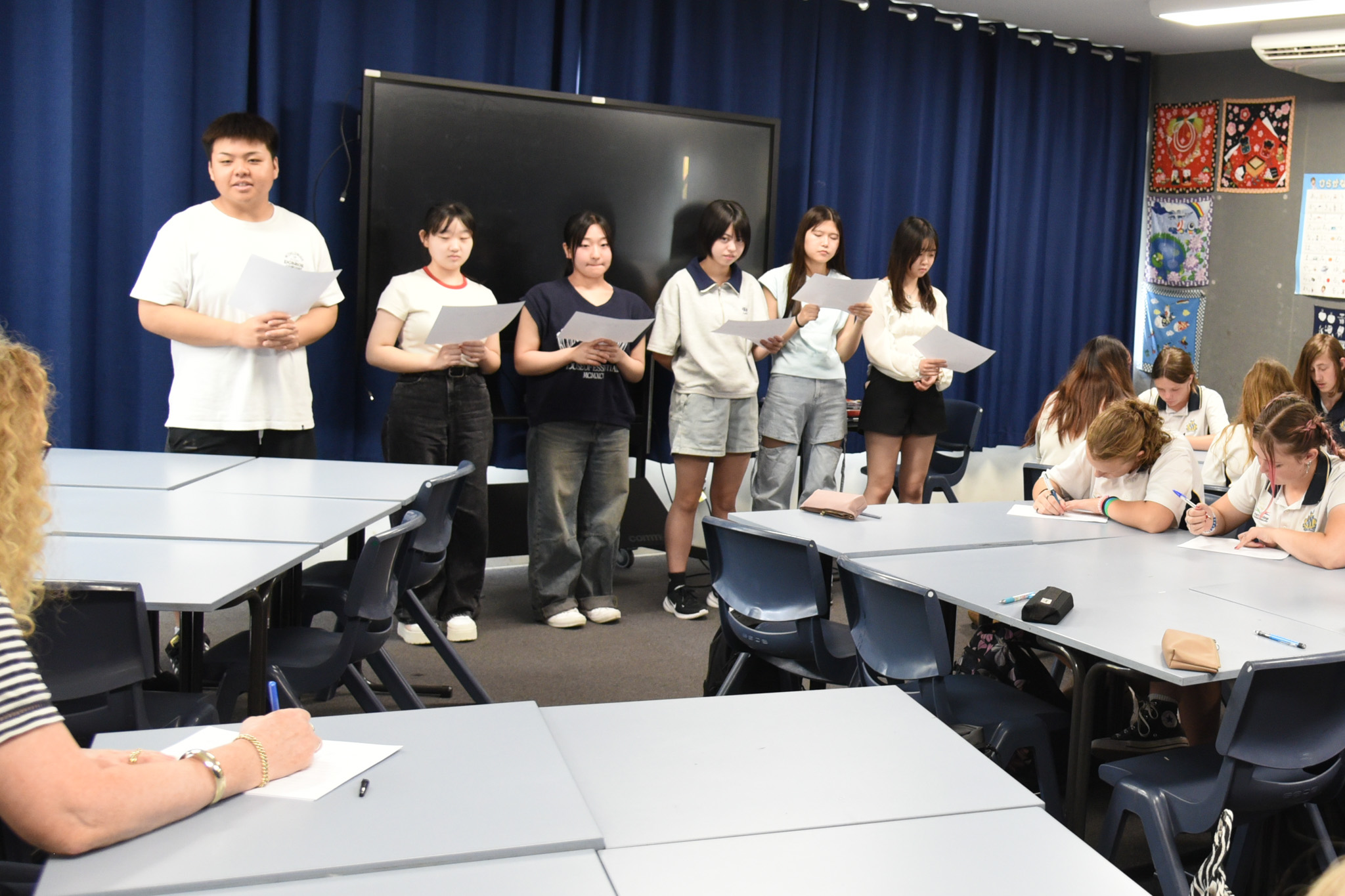 教室の前方に一列に並び、紙を見ながら順番に発表する団員たちの様子。