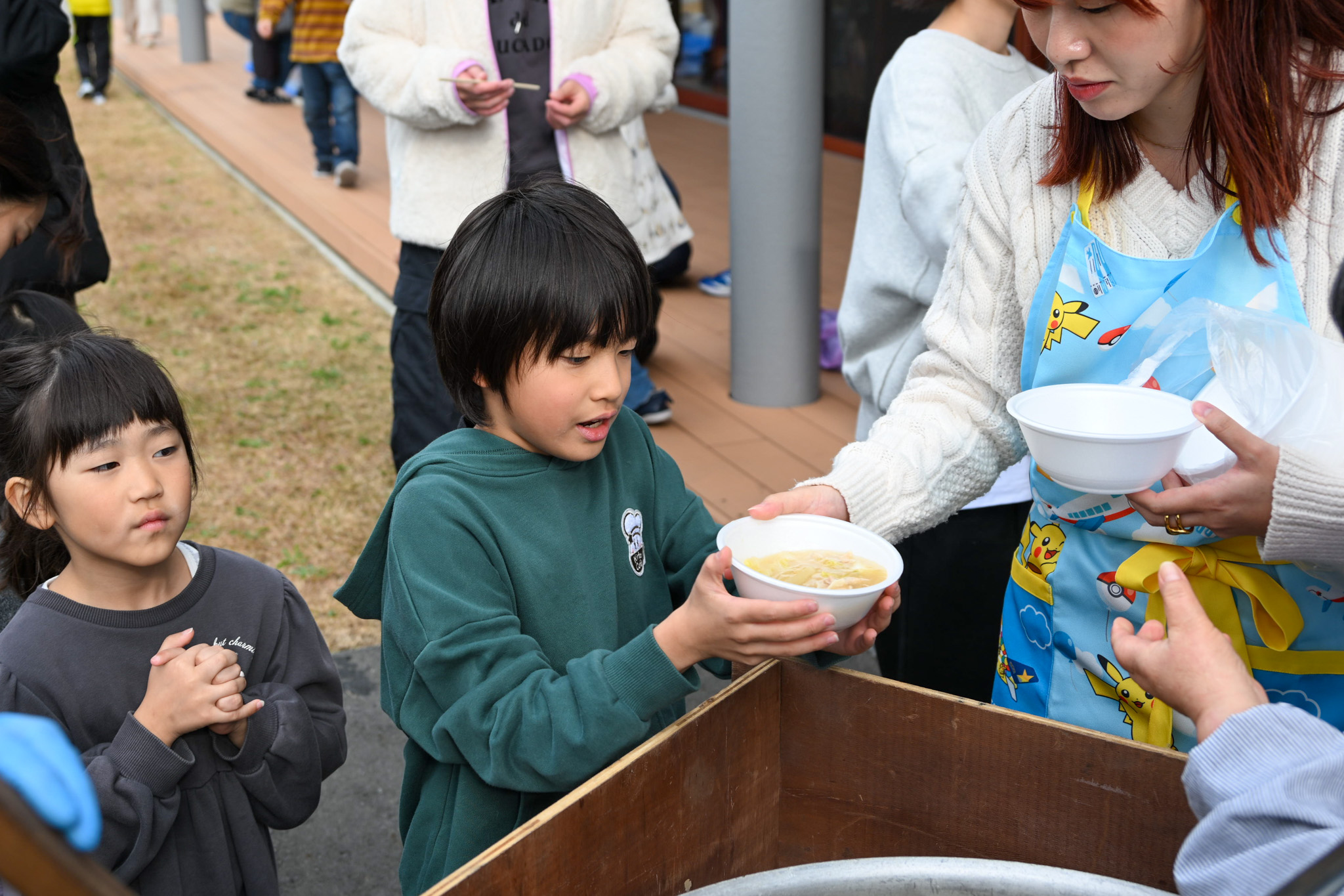 室内の調理室では家庭用の鍋で子どもたちが豚汁を作り、屋外では地域のボランティアの皆さんが大鍋で豚汁を作りました。写真は、外の大鍋で作った豚汁を受け取る様子です。
