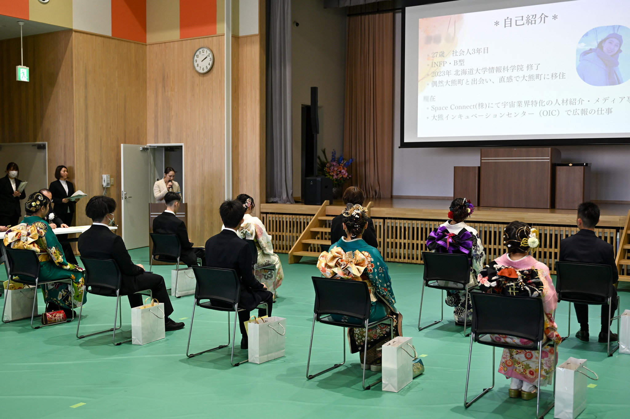 東日本大震災当時は未就学児だった出席者に「大熊町の今」を伝えるため、講演が行われました。講師の斎藤心さん（大熊インキュベーション勤務）は移住の経緯や展望を語り、「楽しく生きるには本気で取り組み、困難を乗り越えることが大切」と伝えました。写真は、スクリーンに映し出されたスライドを見ながら講演に耳を傾ける新成人の様子です。