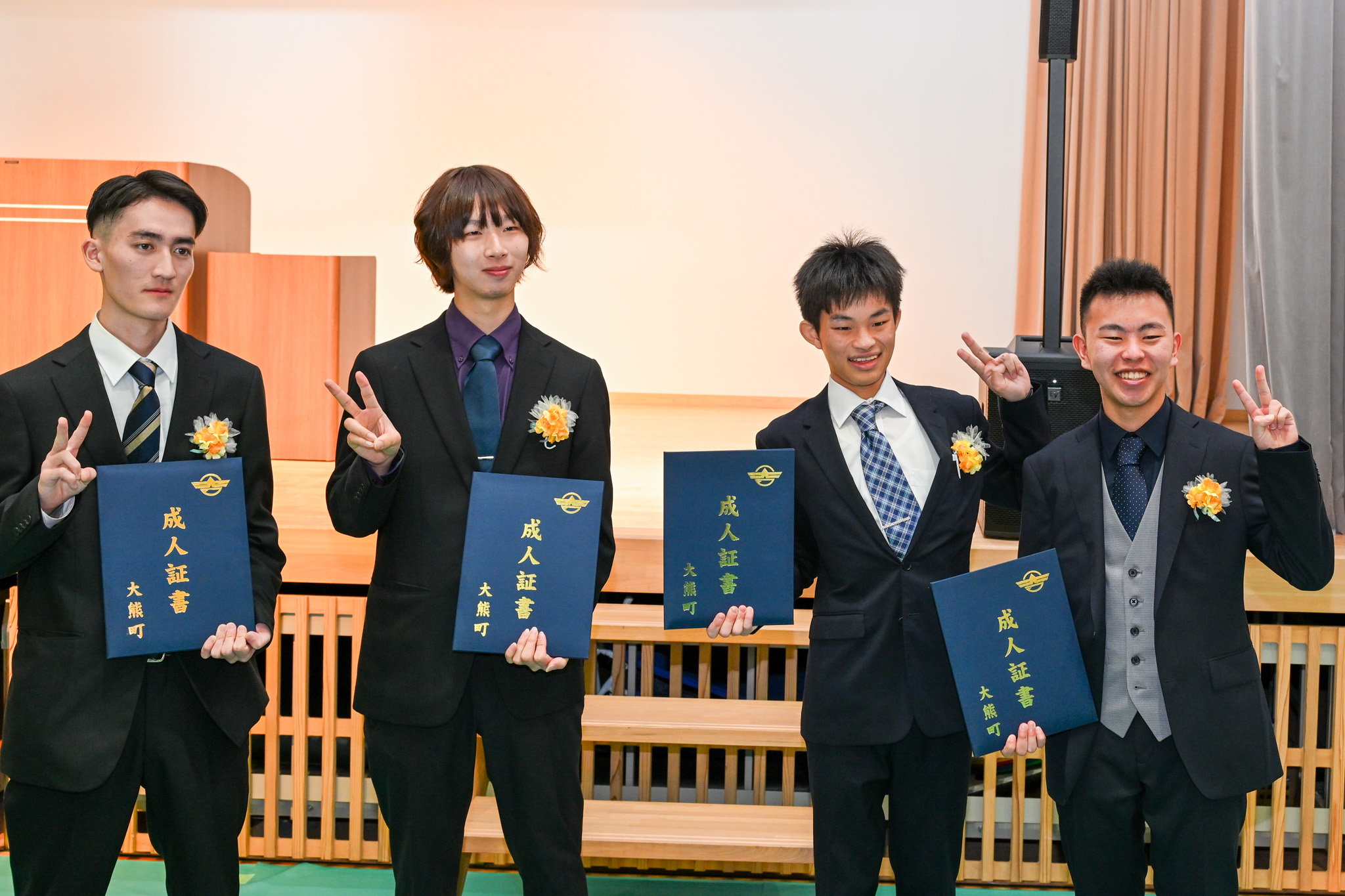 写真は、式後の緊張がほぐれ、成人証書を手にした男性4人がカメラに向かってピースサインを送る楽しそうな様子です。
