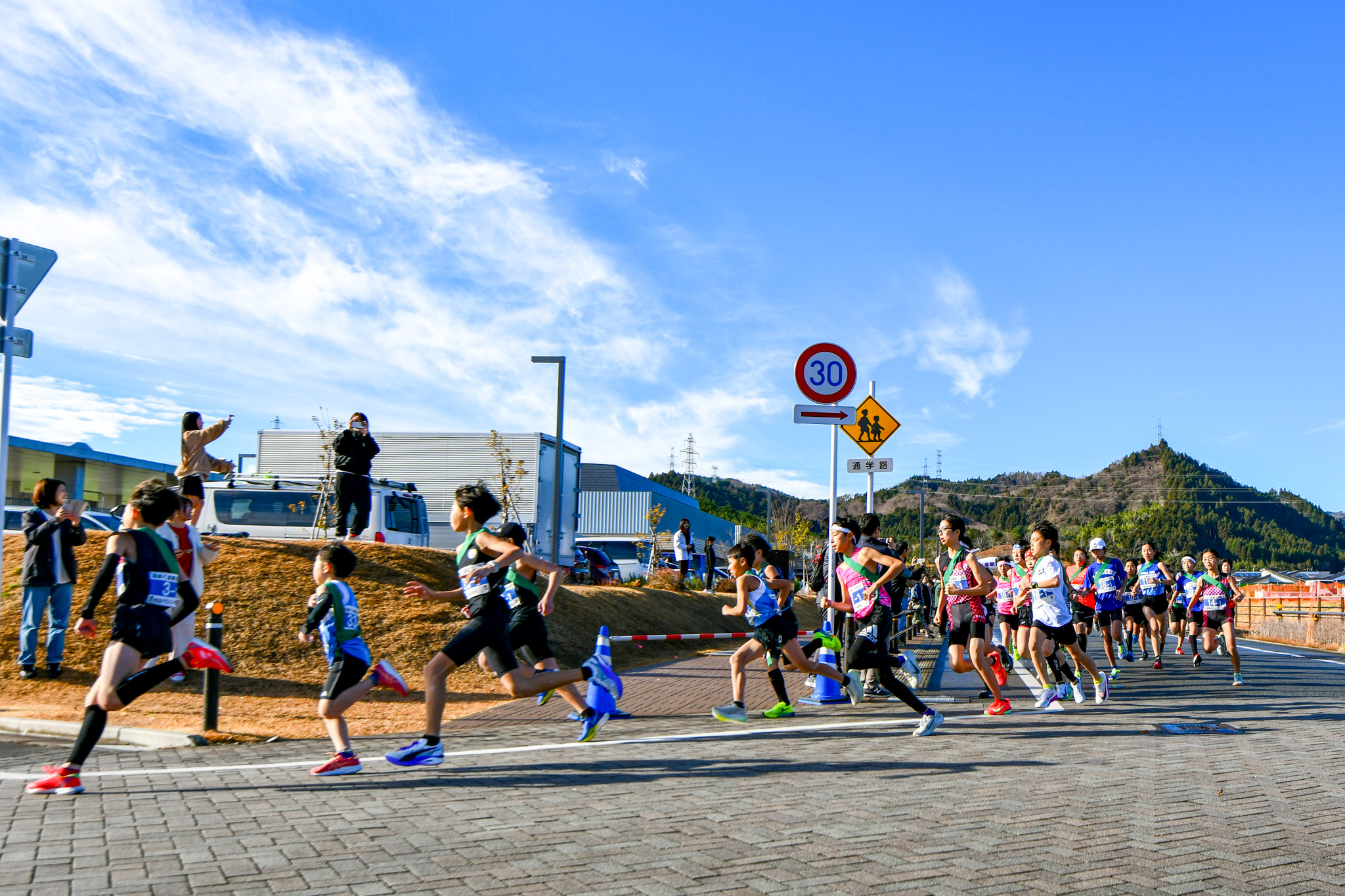 駅伝は学び舎ゆめの森グラウンド前を発着点とする特設コースで行われ、小学生は約1.5km、一般（中学生以上）は約2kmを4人でタスキをつないで走りました。写真は先頭集団に食らいつこうと懸命に走る小学生の選手たちの様子です。